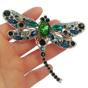May include: A gold-coloured dragonfly brooch, embellished with green, blue, and iridescent crystals. The brooch has a large green central stone and smaller stones outlining the wings and body, creating a sparkling, detailed design. The wings are decorated with various shades of green and blue crystals.