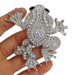 May include: A silver-toned frog brooch, encrusted with clear crystals. The frog has large eyes, webbed feet, and is posed with its arms and legs outstretched. The brooch is a detailed, sparkling accessory.
