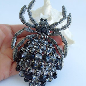 Może przedstawiać: Duża, bogato zdobiona broszka w kształcie pająka. Broszka w kolorze srebrnym ma korpus i nogi inkrustowane czarnymi i szarymi kryształami. Brzuch pająka to skupisko kryształ&oacute;w o r&oacute;żnych rozmiarach. Efektowna biżuteria.