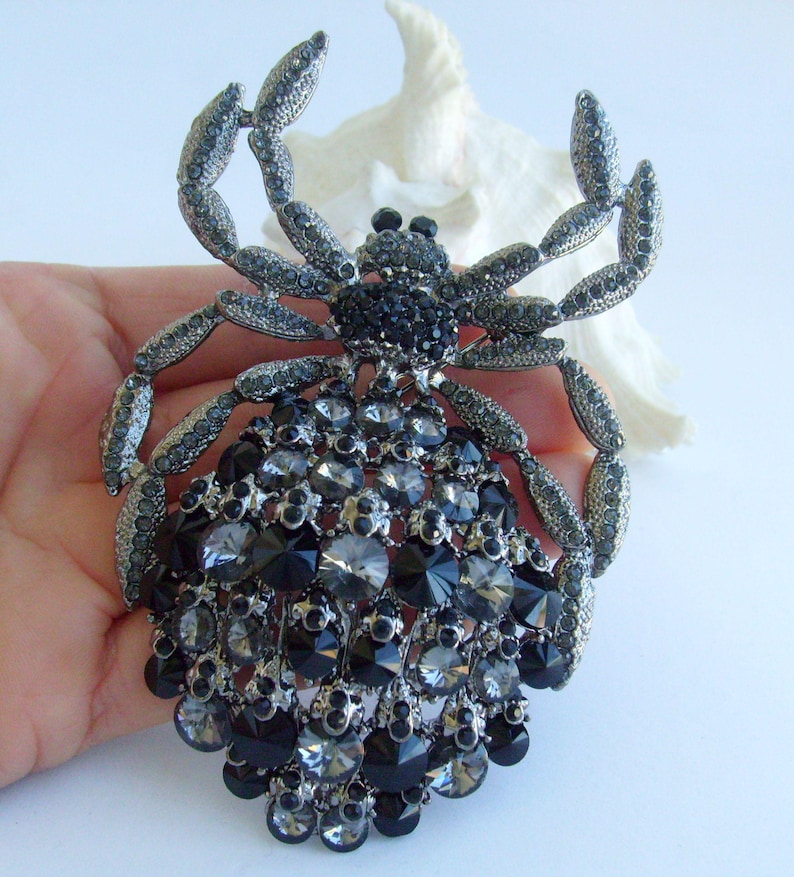 Może przedstawiać: Duża, bogato zdobiona broszka w kształcie pająka. Broszka w kolorze srebrnym ma korpus i nogi inkrustowane czarnymi i szarymi kryształami. Brzuch pająka to skupisko kryształ&oacute;w o r&oacute;żnych rozmiarach. Efektowna biżuteria.