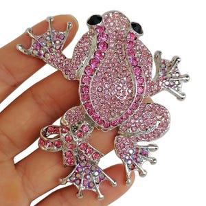 May include: A jeweled frog brooch with a silver-tone metal base. The frog is covered in pink rhinestones, with darker pink stones outlining the body. The eyes are black. The feet have iridescent rhinestones.