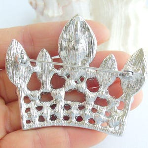 以下が含まれることがあります： テクスチャ加工された表面とピン留めが付いた、銀色の王冠ブローチ。 ブローチは、王冠に似た、オープンなスペースと尖った先端を備えた詳細なデザインが特徴です。