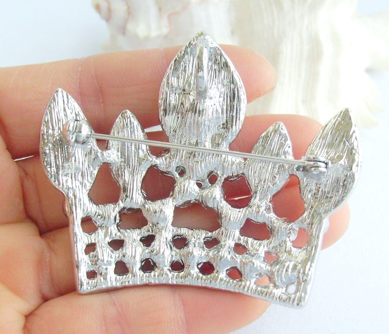 以下が含まれることがあります： テクスチャ加工された表面とピン留めが付いた、銀色の王冠ブローチ。 ブローチは、王冠に似た、オープンなスペースと尖った先端を備えた詳細なデザインが特徴です。