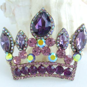 以下が含まれることがあります： ヴィンテージゴールドトーンのメタルフレームに宝石をちりばめた王冠ブローチ。王冠は、大きな涙型の紫色の宝石と、小さく丸い紫と虹色の石が特徴です。気品のあるアクセサリーです。