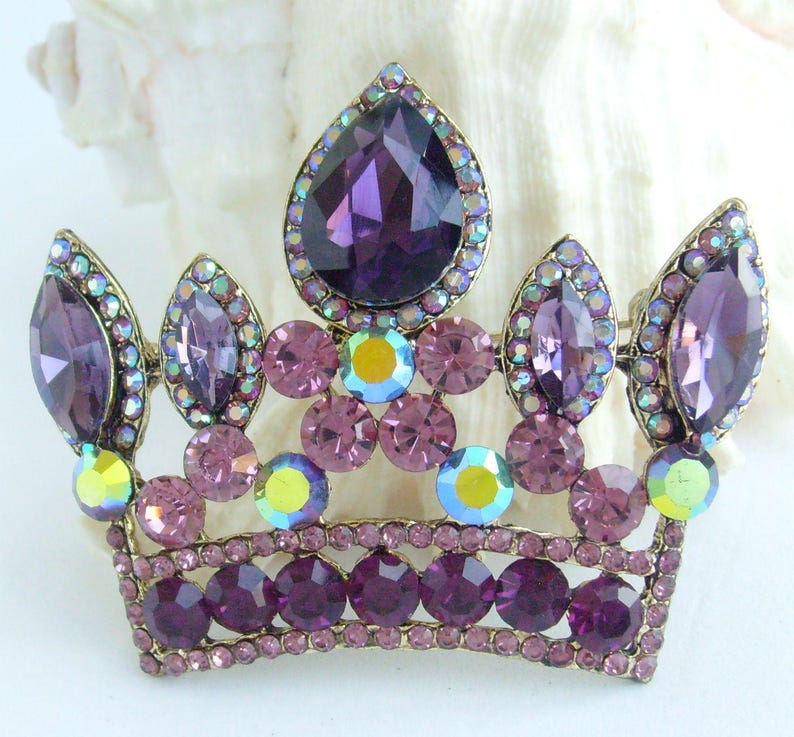 以下が含まれることがあります： ヴィンテージゴールドトーンのメタルフレームに宝石をちりばめた王冠ブローチ。王冠は、大きな涙型の紫色の宝石と、小さく丸い紫と虹色の石が特徴です。気品のあるアクセサリーです。