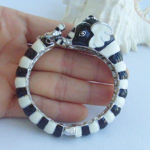 Puede incluir: Una pulsera de brazalete de elefante esmaltado en blanco y negro. La pulsera presenta una banda de metal plateado con segmentos de esmalte blanco y negro alternados. La cabeza del elefante está detallada con orejas blancas y un ojo negro. La pulsera se sostiene en una mano.