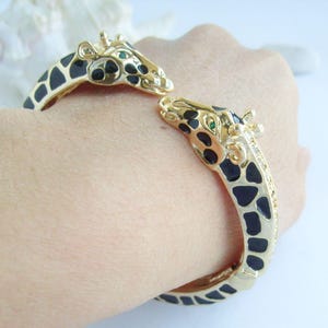 May include: Gold-coloured cuff bracelet featuring giraffe heads with black and cream-coloured spots. The giraffe heads have green eyes and are detailed with black spots. The bracelet is a unique animal-themed accessory.