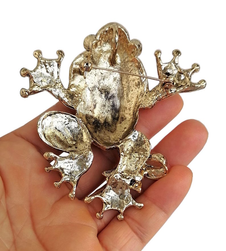 May include: A gold-toned frog brooch with outstretched limbs and textured details. The brooch has a pin on the back for secure attachment. The frog's design is detailed, with a textured surface and a shiny finish, perfect for adding a touch of whimsy to any outfit.