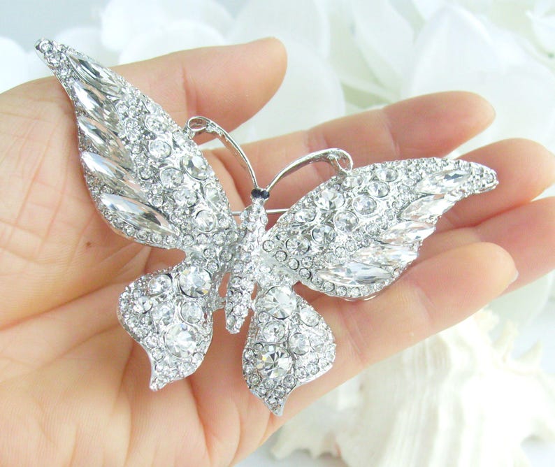May include: A silver butterfly brooch encrusted with clear crystals. The brooch features detailed wings and antennae, with a pin clasp on the back. The butterfly is a sparkling accessory.