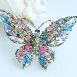 May include: A gold-toned butterfly brooch, encrusted with a variety of colorful gemstones. The brooch features a symmetrical design with wings adorned in shades of pink, blue, green, and clear crystals. The butterfly's body is detailed with smaller, darker gems.