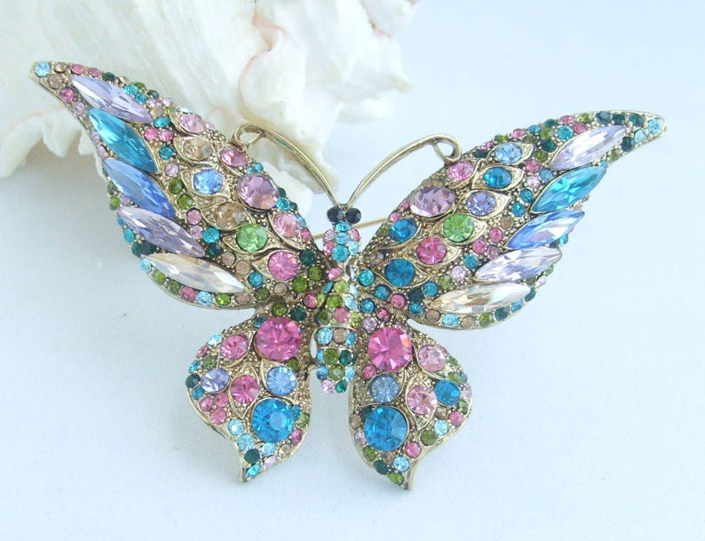 May include: A gold-toned butterfly brooch, encrusted with a variety of colorful gemstones. The brooch features a symmetrical design with wings adorned in shades of pink, blue, green, and clear crystals. The butterfly's body is detailed with smaller, darker gems.