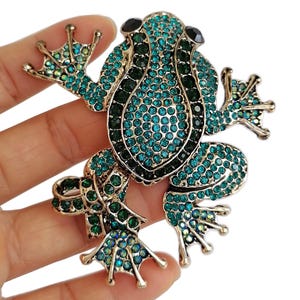 May include: A jeweled frog brooch with a gold-tone metal frame. The frog is covered in teal and dark green rhinestones, with black stones for eyes. The brooch is a detailed, decorative accessory.