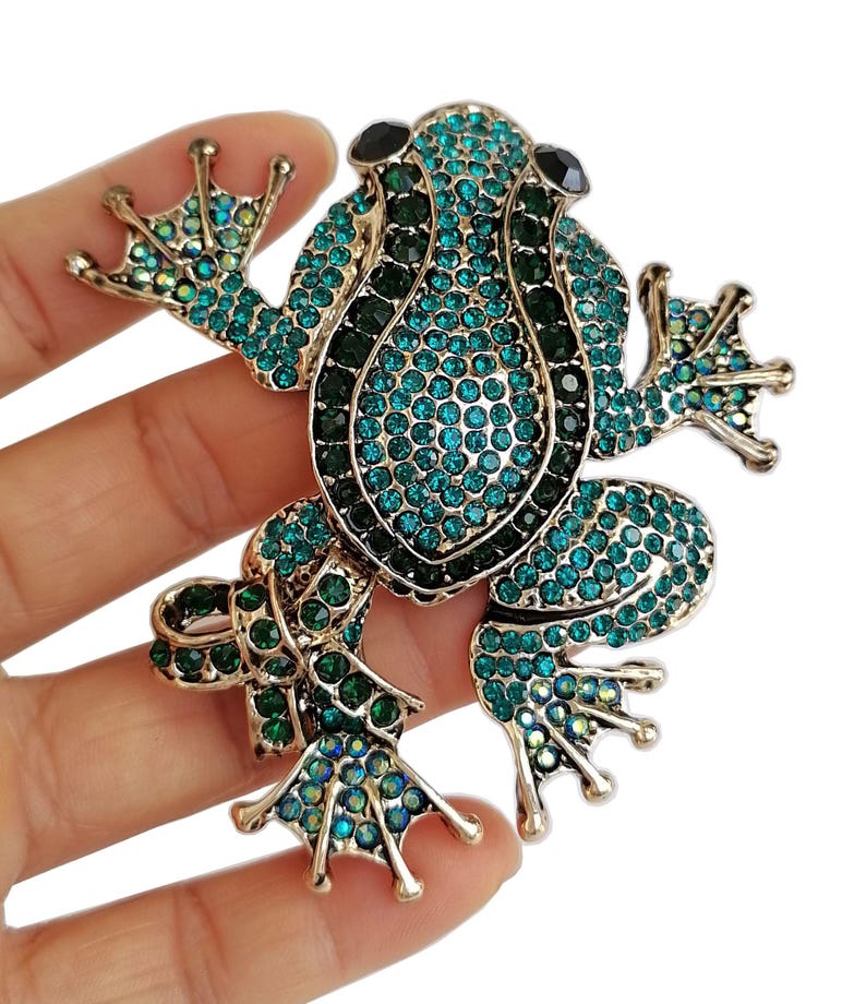 May include: A jeweled frog brooch with a gold-tone metal frame. The frog is covered in teal and dark green rhinestones, with black stones for eyes. The brooch is a detailed, decorative accessory.