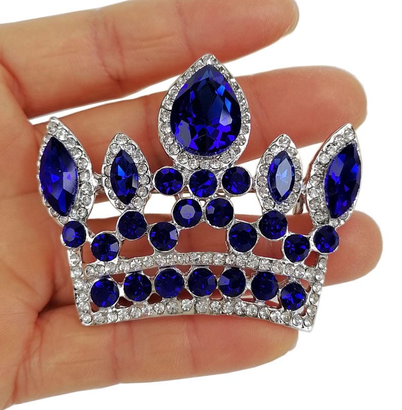 以下が含まれることがあります： 大きな深い青色のティアドロップ型と楕円形の宝石が特徴の、シルバーカラーのクラウンブローチ。小さな透明なクリスタルで囲まれています。クラウンのデザインには、丸い青い宝石の列が含まれています。