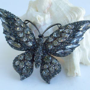 May include: A dark gray butterfly brooch, encrusted with clear and gray crystals. The brooch has a detailed design with a metal frame and is set against a white background.