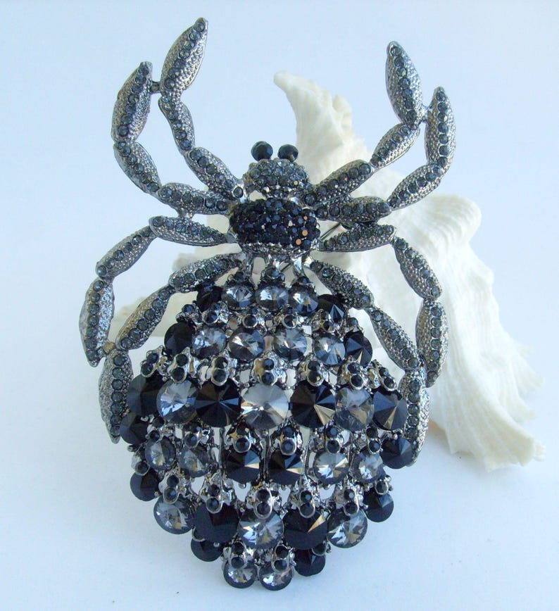 Może przedstawiać: Duża broszka w kształcie kraba. Broszka jest gł&oacute;wnie w kolorze srebrnym i ozdobiona licznymi czarnymi i szarymi kryształami. Ciało i szczypce kraba są szczeg&oacute;łowo wykonane z kryształ&oacute;w.