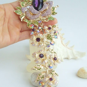 May include: A gold-toned floral brooch with a large purple gemstone center. The brooch features a rose design with lavender and green accents, and dangling floral elements with purple and iridescent crystals.