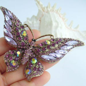 May include: A vintage-style butterfly brooch with a bronze-tone metal frame. The butterfly's wings are adorned with numerous purple and iridescent crystals. The brooch is a decorative accessory.