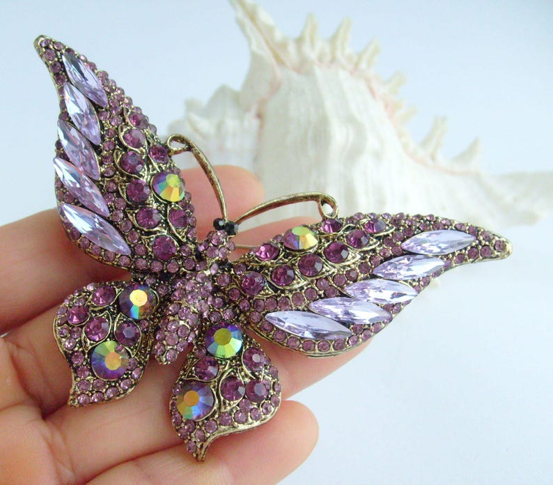 May include: A vintage-style butterfly brooch with a bronze-tone metal frame. The butterfly's wings are adorned with numerous purple and iridescent crystals. The brooch is a decorative accessory.