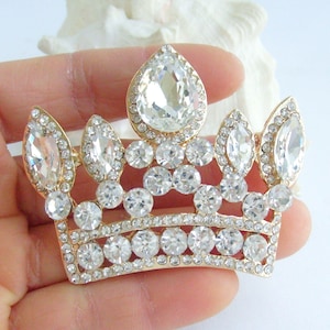 以下が含まれることがあります： 多数の透明で輝くラインストーンで飾られた、金色の王冠ブローチ。ブローチは、上部に大きな洋梨型のラインストーンと、全体に小さな丸いラインストーンが特徴です。王冠はクラシックで堂々とした形をしています。