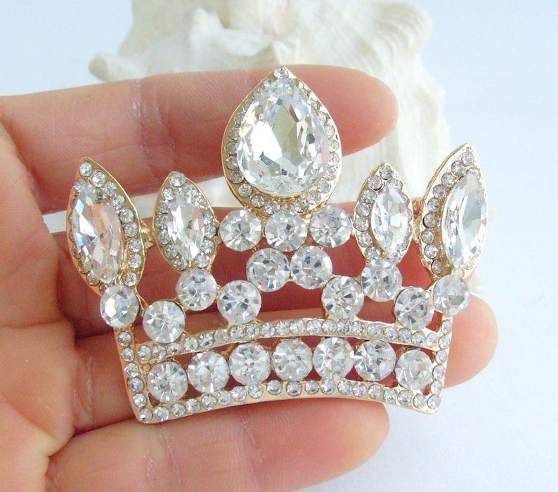 以下が含まれることがあります： 多数の透明で輝くラインストーンで飾られた、金色の王冠ブローチ。ブローチは、上部に大きな洋梨型のラインストーンと、全体に小さな丸いラインストーンが特徴です。王冠はクラシックで堂々とした形をしています。