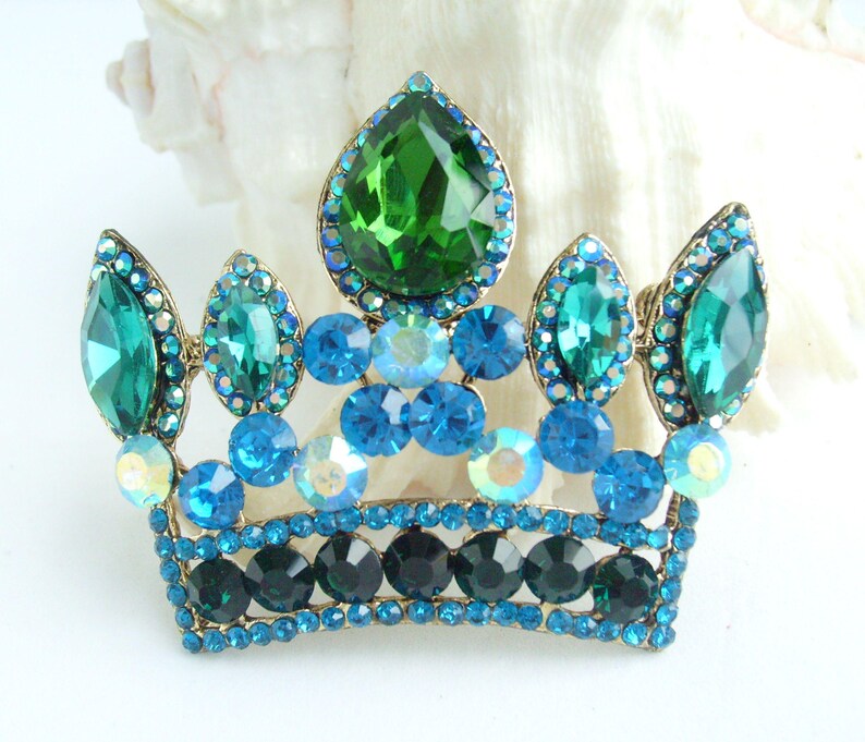 以下が含まれることがあります： 金色の金属フレームに宝石をちりばめた王冠のブローチ。王冠は、大きな涙型の石や小さな丸い石など、さまざまな青と緑のラインストーンで飾られています。ブローチは白い背景に配置されています。