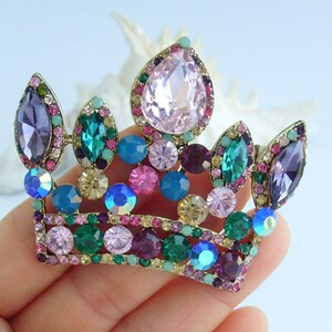 以下が含まれることがあります： 金色の金属ベースに宝石をちりばめた王冠のブローチ。王冠は、ピンク、紫、青、緑、クリアクリスタルなど、さまざまなカラフルな宝石で飾られています。