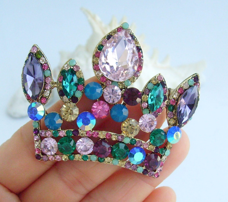 以下が含まれることがあります： 金色の金属ベースに宝石をちりばめた王冠のブローチ。王冠は、ピンク、紫、青、緑、クリアクリスタルなど、さまざまなカラフルな宝石で飾られています。