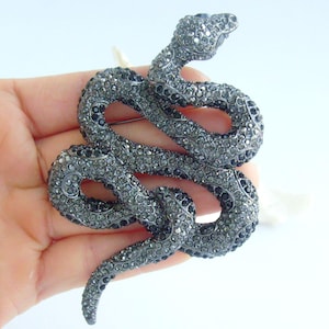 May include: A large, coiled snake brooch. The snake is covered in small, dark gray and black crystals. The brooch has a pin on the back for attachment. The snake's head is raised, and its body is curved into a series of loops.