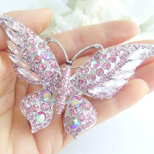 May include: A silver-toned butterfly brooch with pink and iridescent crystals. The brooch features a detailed design with a symmetrical wing pattern. The crystals are round and marquise-shaped, adding sparkle and dimension to the accessory.