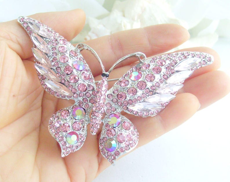 May include: A silver-toned butterfly brooch with pink and iridescent crystals. The brooch features a detailed design with a symmetrical wing pattern. The crystals are round and marquise-shaped, adding sparkle and dimension to the accessory.