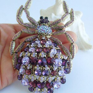 Może przedstawiać: Ozdobna broszka w kształcie kraba lub pająka. Broszka ma złotą metalową ramę i jest bogato zdobiona kryształami w kolorze fioletowym i lawendowym o r&oacute;żnych rozmiarach. Środek ma skupisko iryzujących kryształ&oacute;w. Całość jest skomplikowana i przyciąga wzrok.