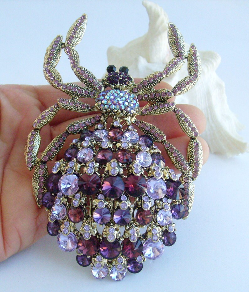 Może przedstawiać: Ozdobna broszka w kształcie kraba lub pająka. Broszka ma złotą metalową ramę i jest bogato zdobiona kryształami w kolorze fioletowym i lawendowym o r&oacute;żnych rozmiarach. Środek ma skupisko iryzujących kryształ&oacute;w. Całość jest skomplikowana i przyciąga wzrok.