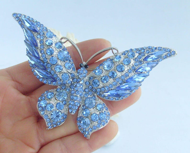 May include: A large, ornate butterfly brooch with light blue crystals. The wings are detailed with various sizes and shapes of blue stones, set in a silver-tone metal. The brooch is designed to be worn as a decorative accessory.