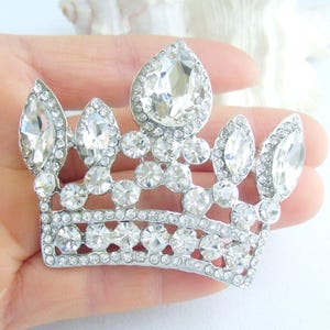 以下が含まれることがあります： 多数の透明で輝くラインストーンがちりばめられた、銀色の王冠ブローチ。王冠は、中央に涙型の石と、複数の小さな丸い石があり、まばゆい効果を生み出しています。ブローチは手で持たれており、その複雑なデザインが示されています。