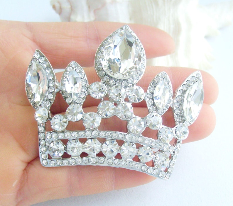 以下が含まれることがあります： 多数の透明で輝くラインストーンがちりばめられた、銀色の王冠ブローチ。王冠は、中央に涙型の石と、複数の小さな丸い石があり、まばゆい効果を生み出しています。ブローチは手で持たれており、その複雑なデザインが示されています。
