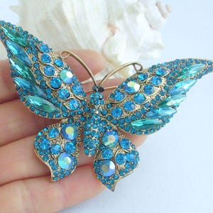 May include: A gold-toned butterfly brooch with turquoise and teal-colored rhinestones. The brooch features a detailed design with various sizes and shapes of sparkling stones, creating a shimmering effect. The butterfly's wings are spread, showcasing the intricate stone arrangement.