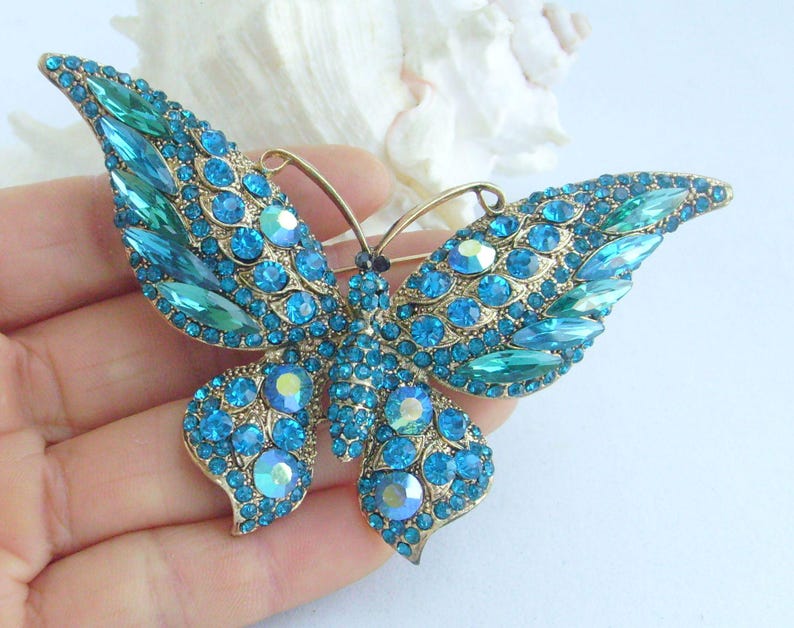 May include: A gold-toned butterfly brooch with turquoise and teal-colored rhinestones. The brooch features a detailed design with various sizes and shapes of sparkling stones, creating a shimmering effect. The butterfly's wings are spread, showcasing the intricate stone arrangement.