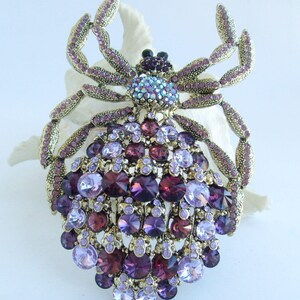 Może przedstawiać: Ozdobna broszka w kształcie pająka. Broszka ma złotą metalową ramę i jest wysadzana r&oacute;żnymi fioletowymi i fioletowymi kryształkami. Ciało pająka składa się z dużych, okrągłych kamieni, a jego nogi są przegubowe.