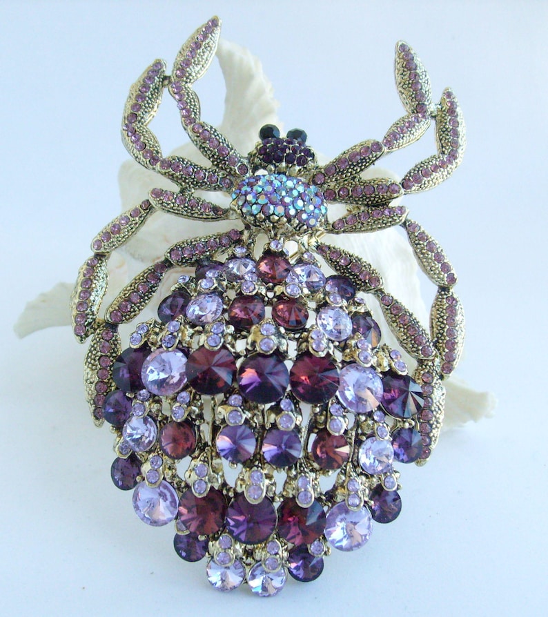 Może przedstawiać: Ozdobna broszka w kształcie pająka. Broszka ma złotą metalową ramę i jest wysadzana r&oacute;żnymi fioletowymi i fioletowymi kryształkami. Ciało pająka składa się z dużych, okrągłych kamieni, a jego nogi są przegubowe.