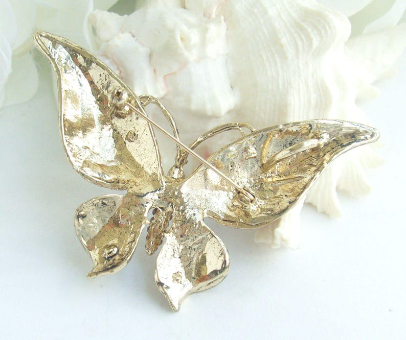 May include: A gold-toned butterfly brooch with textured wings and a pin closure. The butterfly design features detailed veining and a three-dimensional appearance. The brooch is likely made of metal and is suitable for adding to clothing or accessories.