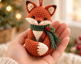 May include: A small, handcrafted crochet fox ornament featuring orange, white, and dark brown yarn. The fox wears a textured green scarf adorned with a small sprig of holly with three red berries. A matching orange yarn loop is attached to the top.