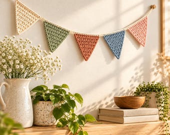 May include: A crochet bunting banner featuring five triangular flags in cream, sage green, terracotta, dusty blue, and soft pink, all connected by a cream-colored yarn string and suspended horizontally against a plain wall.