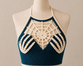 May include: A dark teal crochet halter top featuring a cream-colored spiderweb design across the chest and ribcage area. The garment includes two cream-colored skeletal hand motifs positioned over the bust, with a solid dark teal base.