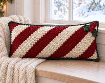 May include: A rectangular crochet pillow featuring diagonal stripes in deep red and cream white. The edges are finished with a dark green border, and the top right corner is decorated with a crocheted holly leaf and berry applique.