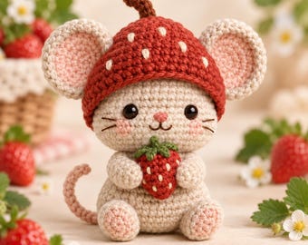 May include: A beige crocheted mouse figurine wearing a red strawberry-shaped hat with white seed details. The mouse has large round ears, pink cheeks, and holds a small red crocheted strawberry in its front paws. It has a curled tail.