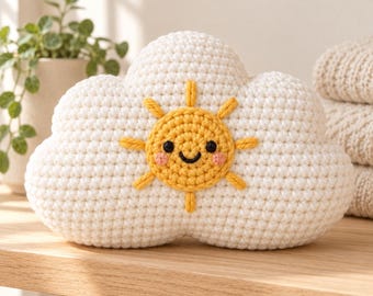 May include: A white crochet cloud pillow featuring a centered yellow sun with a smiling face, black embroidered eyes, and small pink cheeks. The sun has eight radiating yellow rays stitched onto the textured white yarn surface.