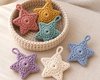 May include: A collection of six crocheted star ornaments in various colors including mustard yellow, dusty rose, sage green, lavender, cream, and periwinkle blue, with three stars resting inside a round, beige crocheted basket.