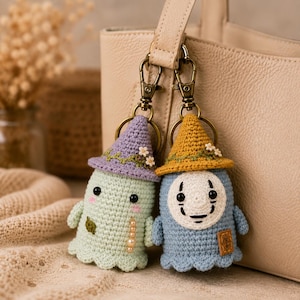 May include: Two crocheted ghost keychains with bronze-toned lobster clasps. The left ghost is light green with a purple hat, small white flowers, and pearl accents. The right ghost is blue with a yellow hat, white flowers, and a white mask face.