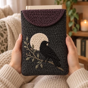 Pode incluir: Uma capa de crochê cinza-escura com fecho de aba em um tom ameixa profundo. A parte frontal apresenta um design bordado de um corvo preto pousado em um galho contra uma grande lua circular de cor creme, feita com pontos detalhados.